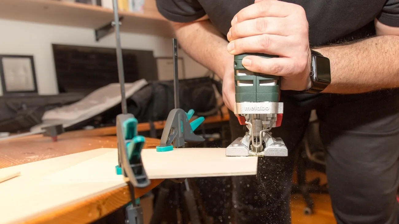 Eine Person sägt eine an einem Tischende festgemachte Holzplatte mit der Metabo Stichsäge.
